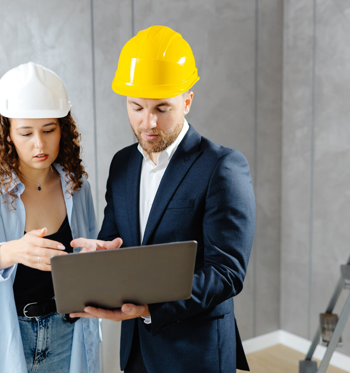 Attraktive junge Bauunternehmerin spricht mit einem Kunden in einem Helm und demonstriert auf einem Laptop die Abfolge von Reparaturen im Wohnzimmer.