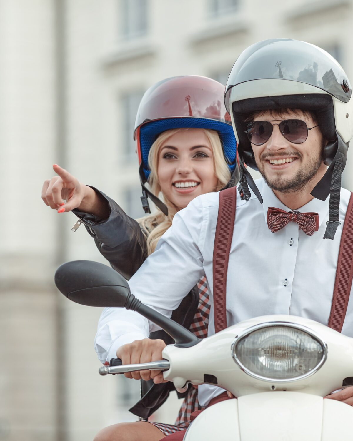 Ein junges Paar in modischer Kleidung und mit Helmen auf dem Kopf sitzt auf einem weißen Motorroller. Der Mann trägt ein weißes Hemd, eine rote Fliege und Hosenträger, während die Frau ein schwarz-weiß kariertes Outfit mit einem blauen Helm trägt. Sie lächelt und zeigt etwas mit dem Finger, während er den Roller lenkt und durch die Straßen der Stadt navigiert