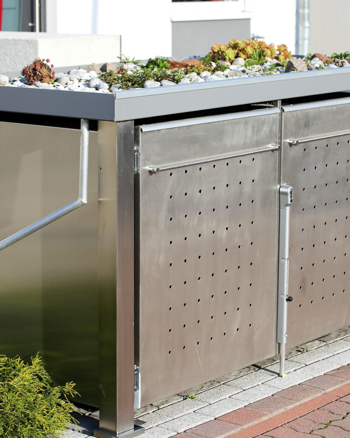Eine moderne Mülltonnenbox aus Edelstahl mit einem bepflanzten Dach steht auf einem gepflasterten Bereich im Garten.