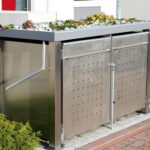 Eine moderne Mülltonnenbox aus Edelstahl mit einem bepflanzten Dach steht auf einem gepflasterten Bereich im Garten.