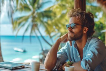 Ein Mann sitzt nachdenklich in einer Strandbar, genießt den Ausblick auf das Meer und die Palmen im Hintergrund. Auf dem Tisch vor ihm liegt ein Notizbuch und eine Tasse Kaffee.