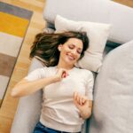 Frau liegt entspannt auf einem Sofa, hört Musik über kabellose Ohrhörer und lächelt, während sie einen entspannten Moment zu Hause genießt.