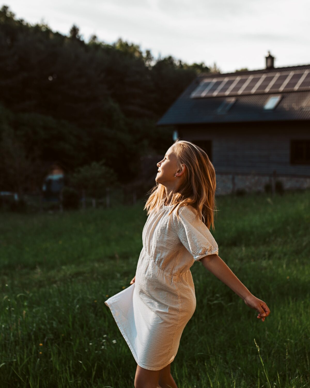 Ein junges Mädchen in einem weißen Kleid steht auf einer Wiese und genießt die Natur. Im Hintergrund sieht man ein modernes Haus mit Photovoltaikanlage auf dem Dach, das Nachhaltigkeit und umweltfreundliches Wohnen symbolisiert