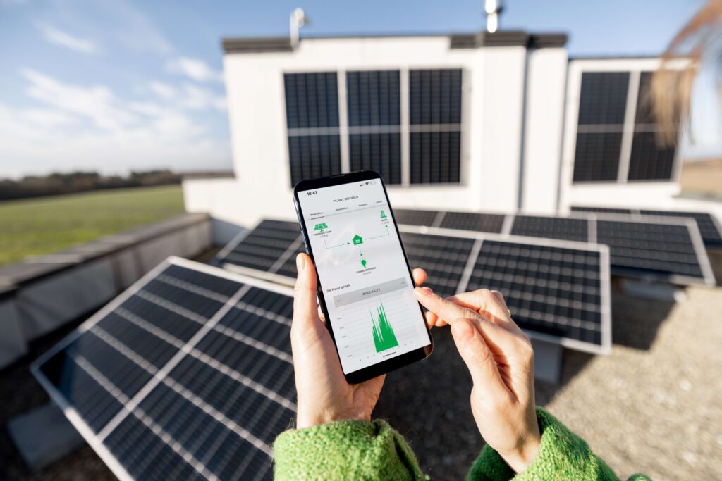 ine Person überwacht die Leistung einer Solaranlage auf ihrem Smartphone, während sie neben den Solarpanels steht. Die Anzeige auf dem Bildschirm zeigt die Energieerzeugung und den Verbrauch, symbolisiert die intelligente Steuerung von erneuerbaren Energien durch einen Fronius Wechselrichter