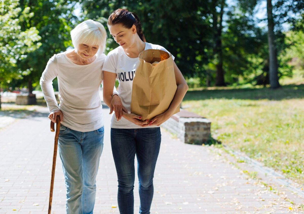 Eine junge Frau begleitet eine ältere Dame mit Gehstock und Einkaufstüte – ein Beispiel für die Aufgaben in der Betreuungsassistent Ausbildung.