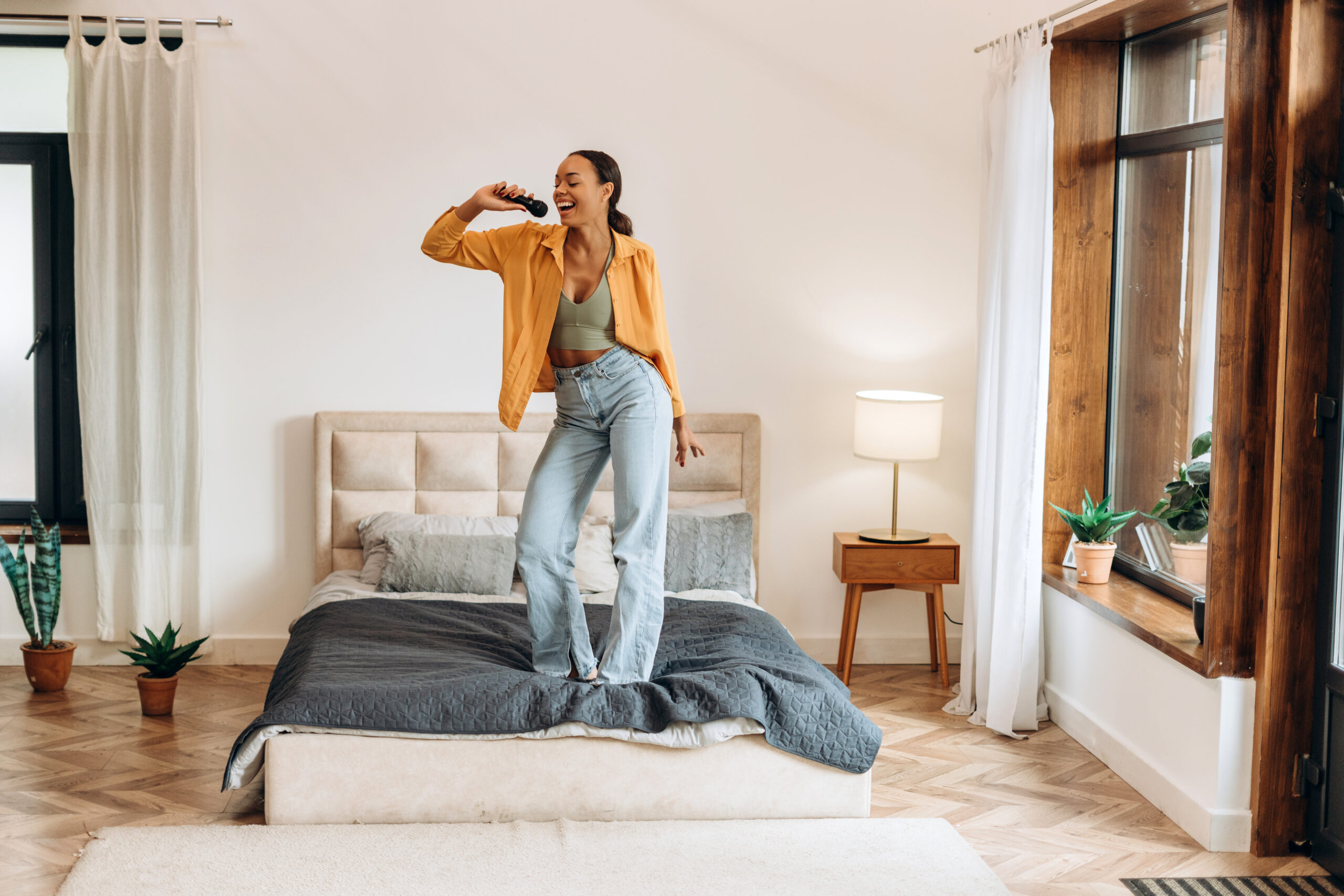 Junge Frau tanzt fröhlich auf einem modernen Polsterbett in einem hellen Schlafzimmer mit Holzfenster – mit spannbettlaken 160x200 in Beige-Grau.